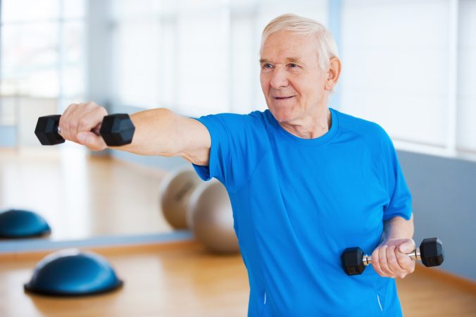 Struggling with age. Confident senior man exercising with dumbbells and smiling while standing in health club