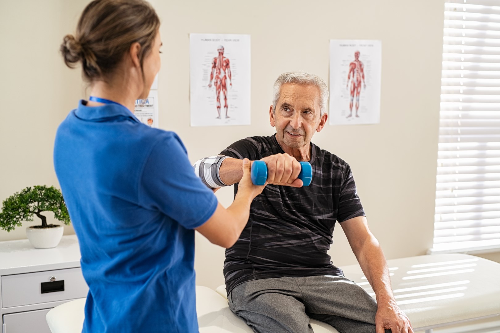 Physiotherapist helping senior man to lift hand weights. Senior man exercising and recovering at nursing home due to epicondylitis. Senior man at care centre exercising with professional physiotherapist.